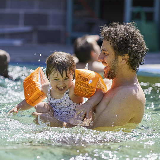 Father holding their toddler wearing orange floatation aid wings smiling in the mushroom pool